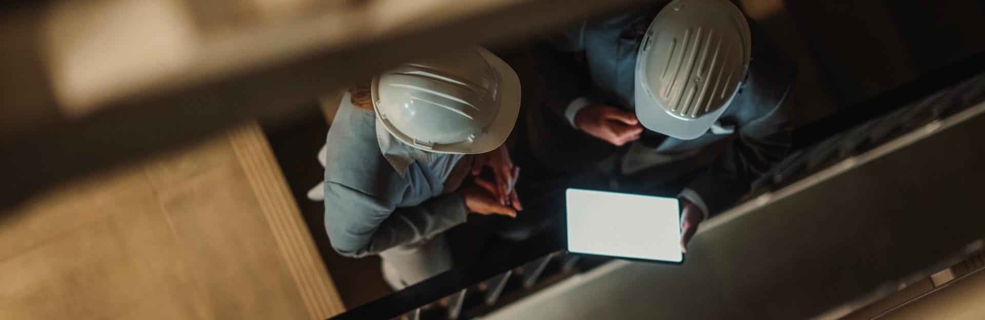 Blick aus einem oberen Stockwerk eines Treppenhauses auf zwei Personen in Businesskleidung mit Helm